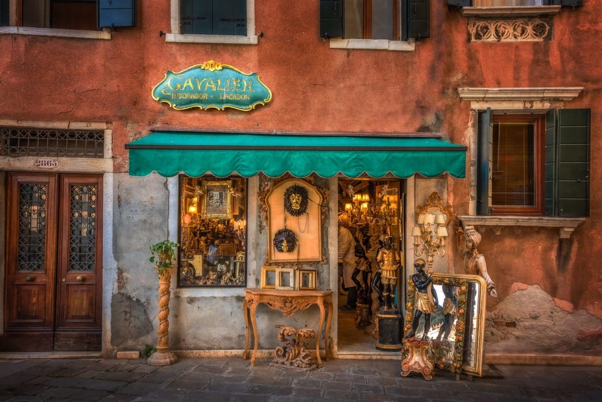 Vintage storefront with green awning, wooden door, and various antique items displayed outside, including mirrors and sculptures.