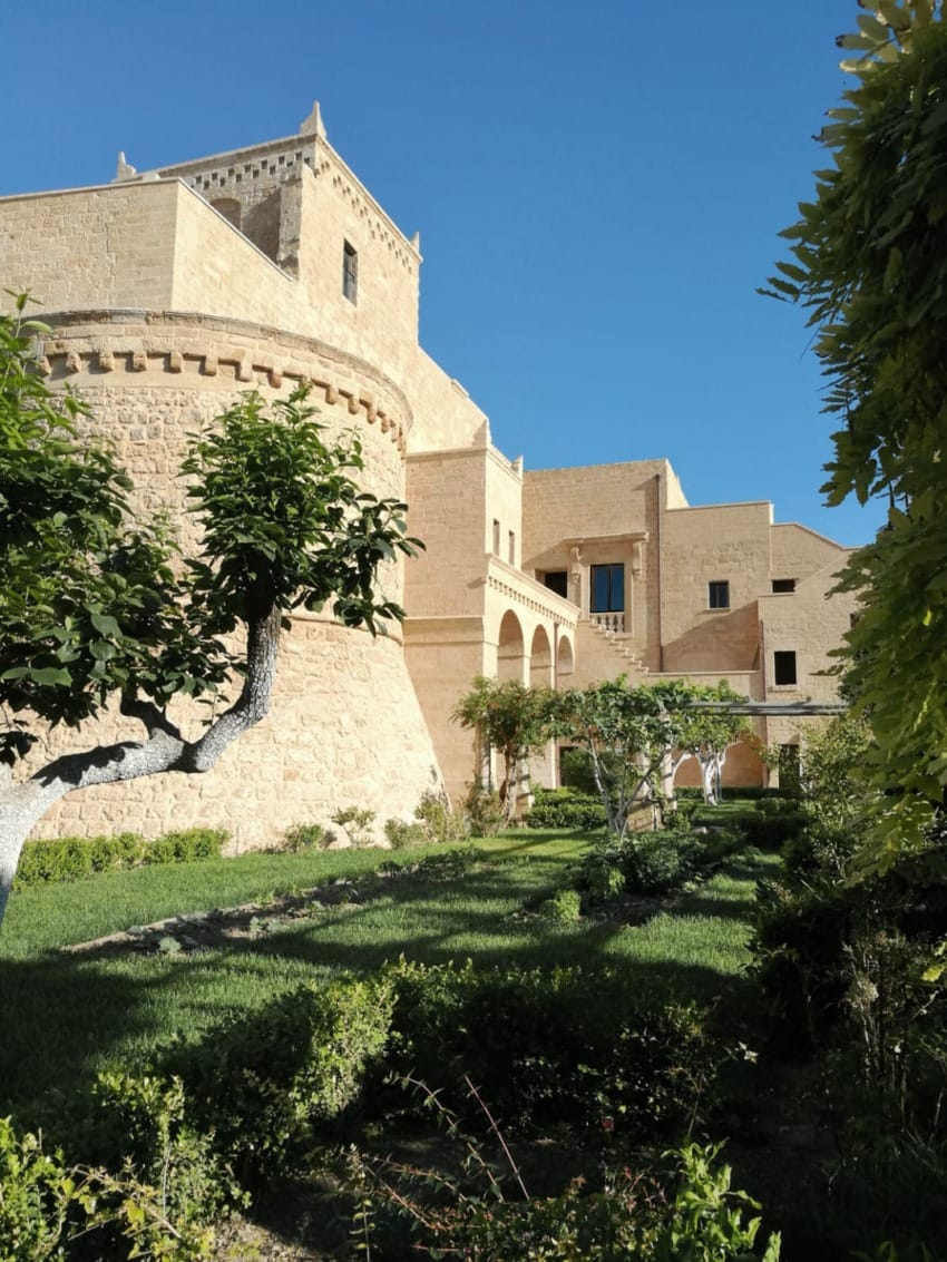 Historic stone fortress with lush garden and blue sky in the background, featuring arched windows and stone walls.