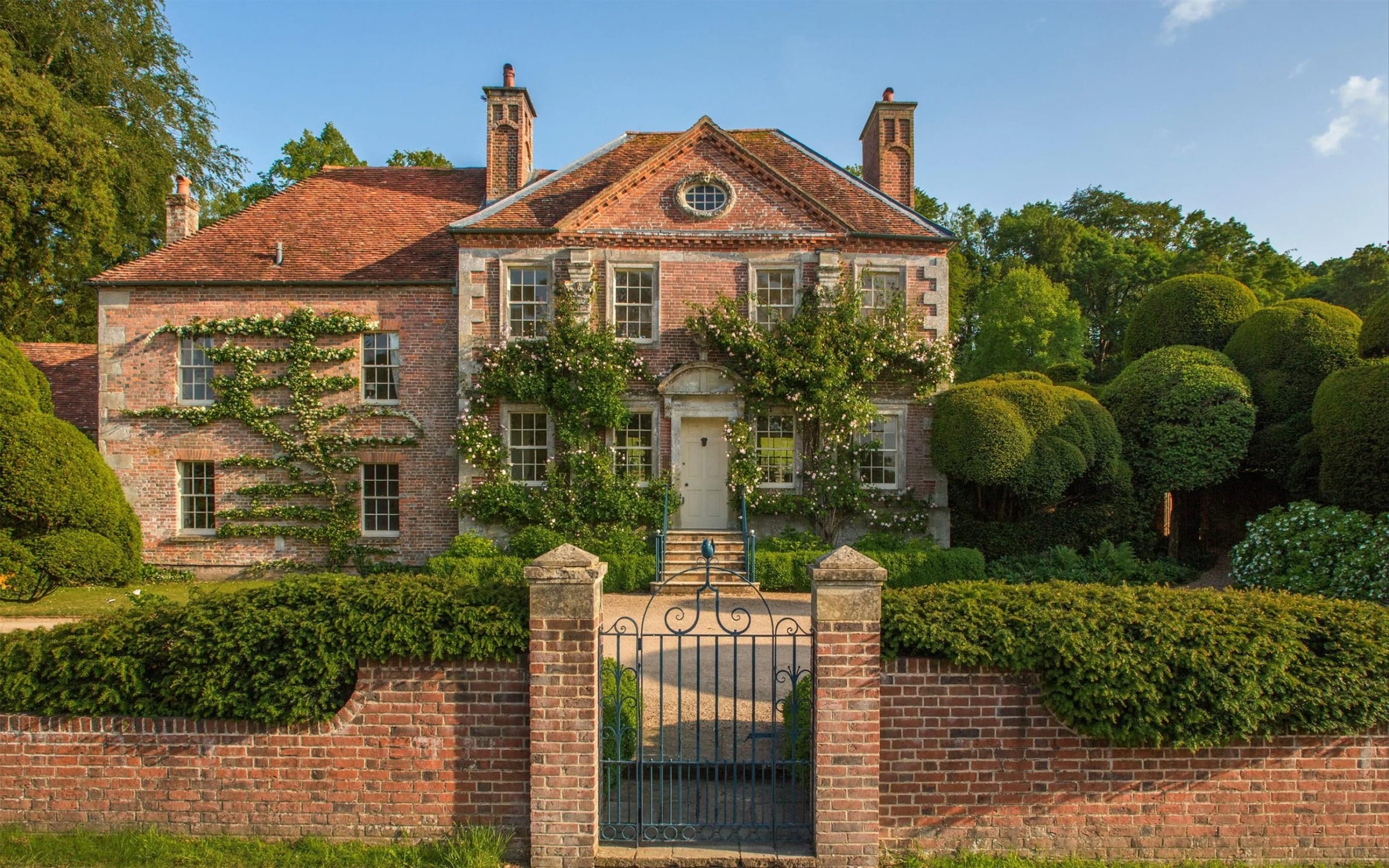 Brick mansion with ivy-covered walls, surrounded by lush gardens and a wrought iron gate, under a clear blue sky.
