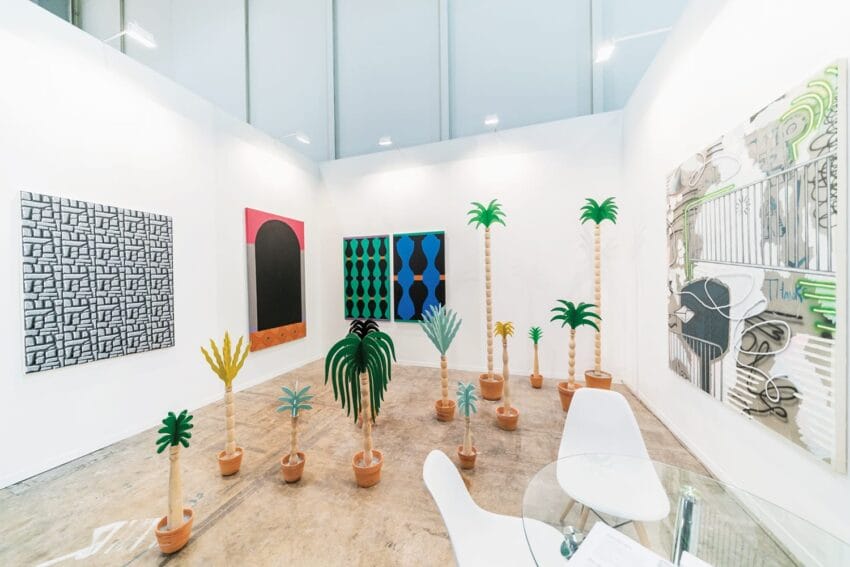 Art gallery with abstract paintings on white walls and a collection of sculpted palm trees in pots on the floor.