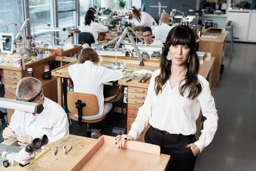 Woman in a professional jewelry workshop with artisans working at benches in the background.