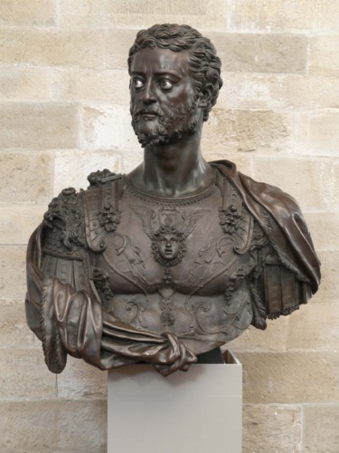 Bronze bust of a bearded man in Roman armor with detailed engravings, displayed against a stone wall background.