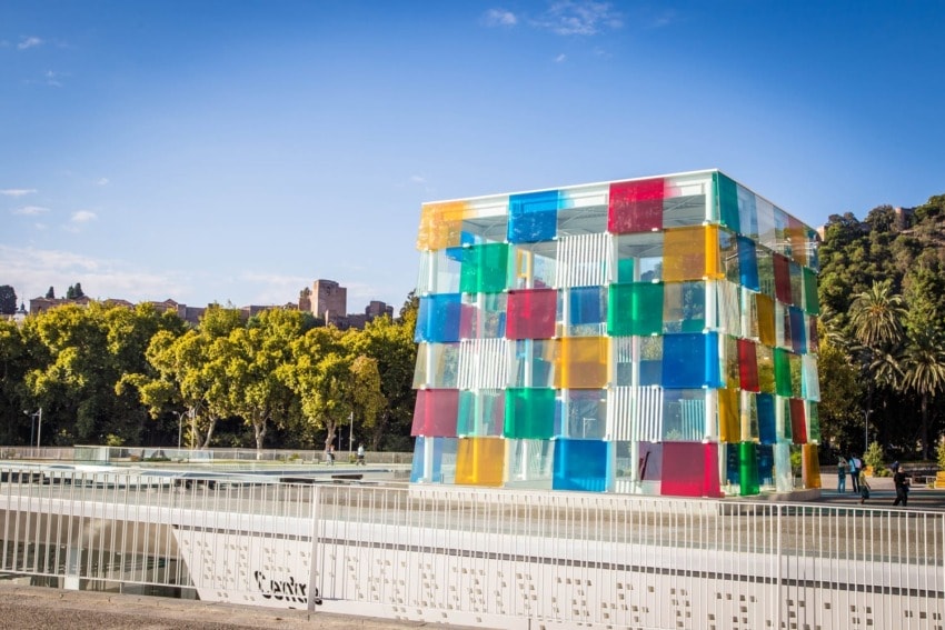 Colorful glass cube structure with vibrant squares, set against a backdrop of trees and clear blue sky.