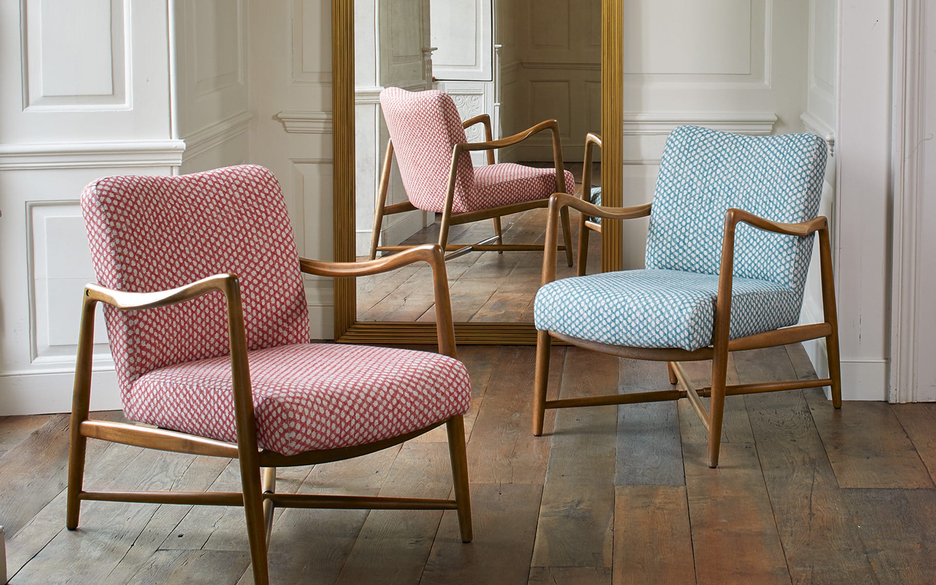 Two upholstered chairs with wooden frames, one red and one blue, placed on a wooden floor in front of a large mirror.