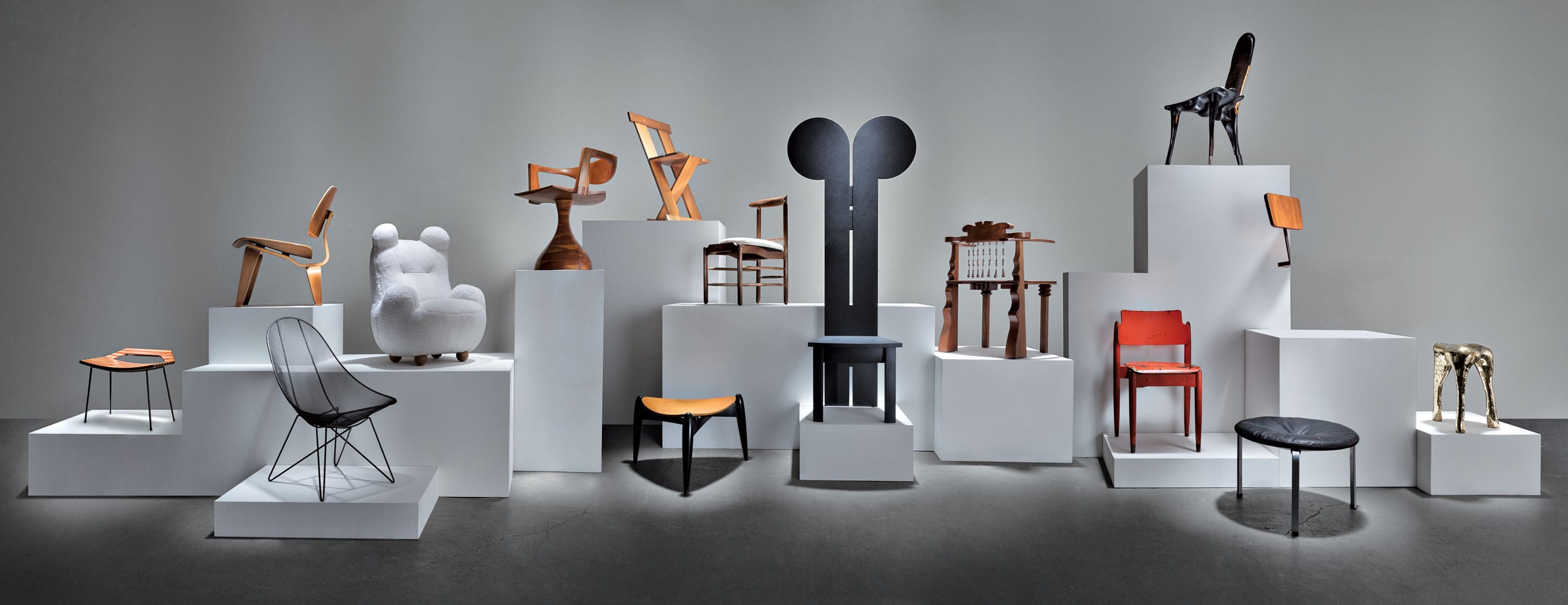 A diverse collection of artistic chairs displayed on a geometric white podium against a gray background.
