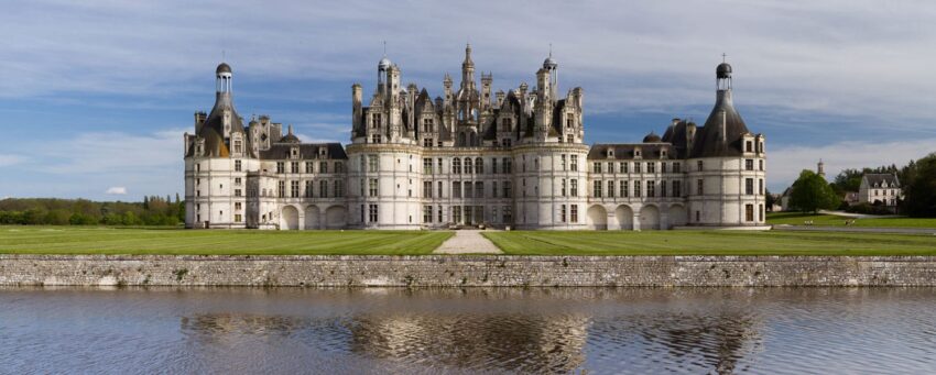 Château de Chambord with manicured lawns and reflection in the water under a partly cloudy sky.
