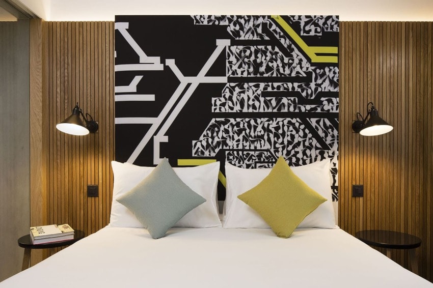Modern hotel room with abstract art, wooden paneling, two wall lamps, and colorful cushions on a neatly made bed.