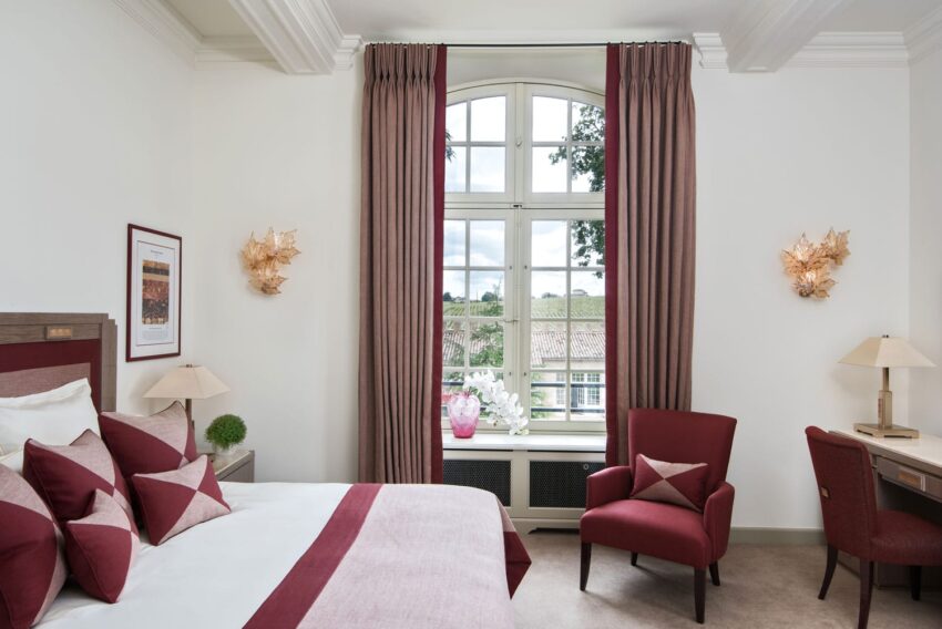Elegant hotel room with red accents, a bed, matching chairs, a window with curtains, and decorative wall lights.