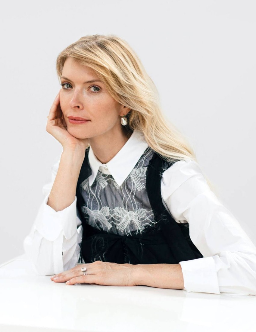 Woman with long blonde hair, wearing a white blouse and black dress, resting her chin on her hand at a table.