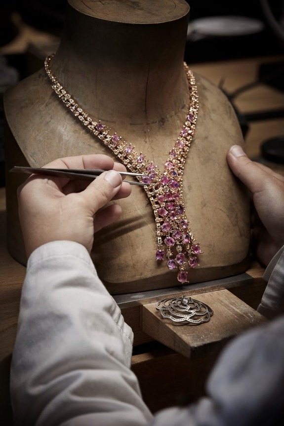 Jeweler meticulously crafting an intricate gold and pink gemstone necklace on a wooden bust in a workshop setting.