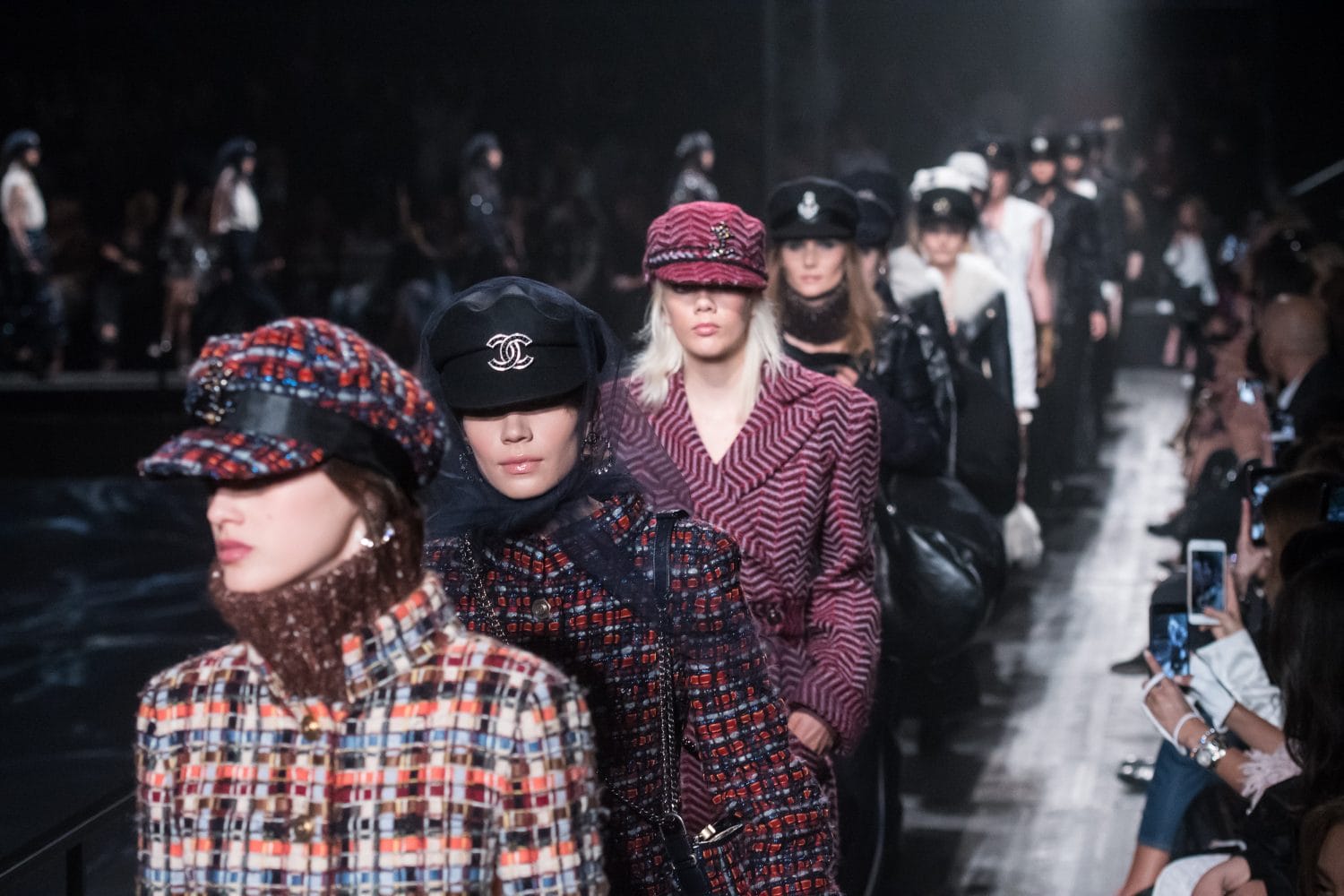 Fashion runway show featuring models in stylish hats and patterned outfits walking down the catwalk.