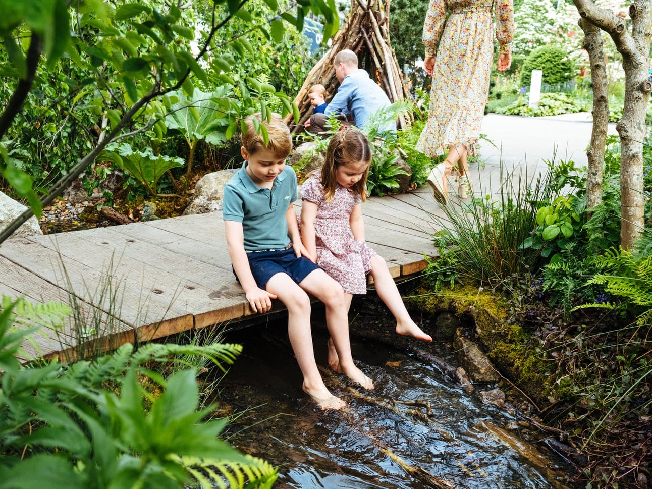 Children sitting on a wooden bridge dipping their feet in a stream surrounded by lush greenery in a garden setting.