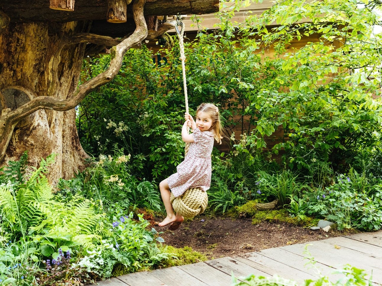 Young girl in a dress happily swinging on a rope swing in a lush green garden.