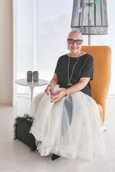 Elderly woman in a black top and white tulle skirt sitting on an orange chair, smiling with hands resting on her lap.