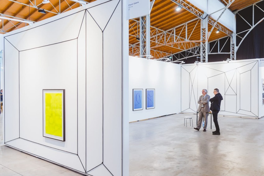 Art gallery with geometric white walls, vibrant yellow painting, and two people standing and observing the artwork.