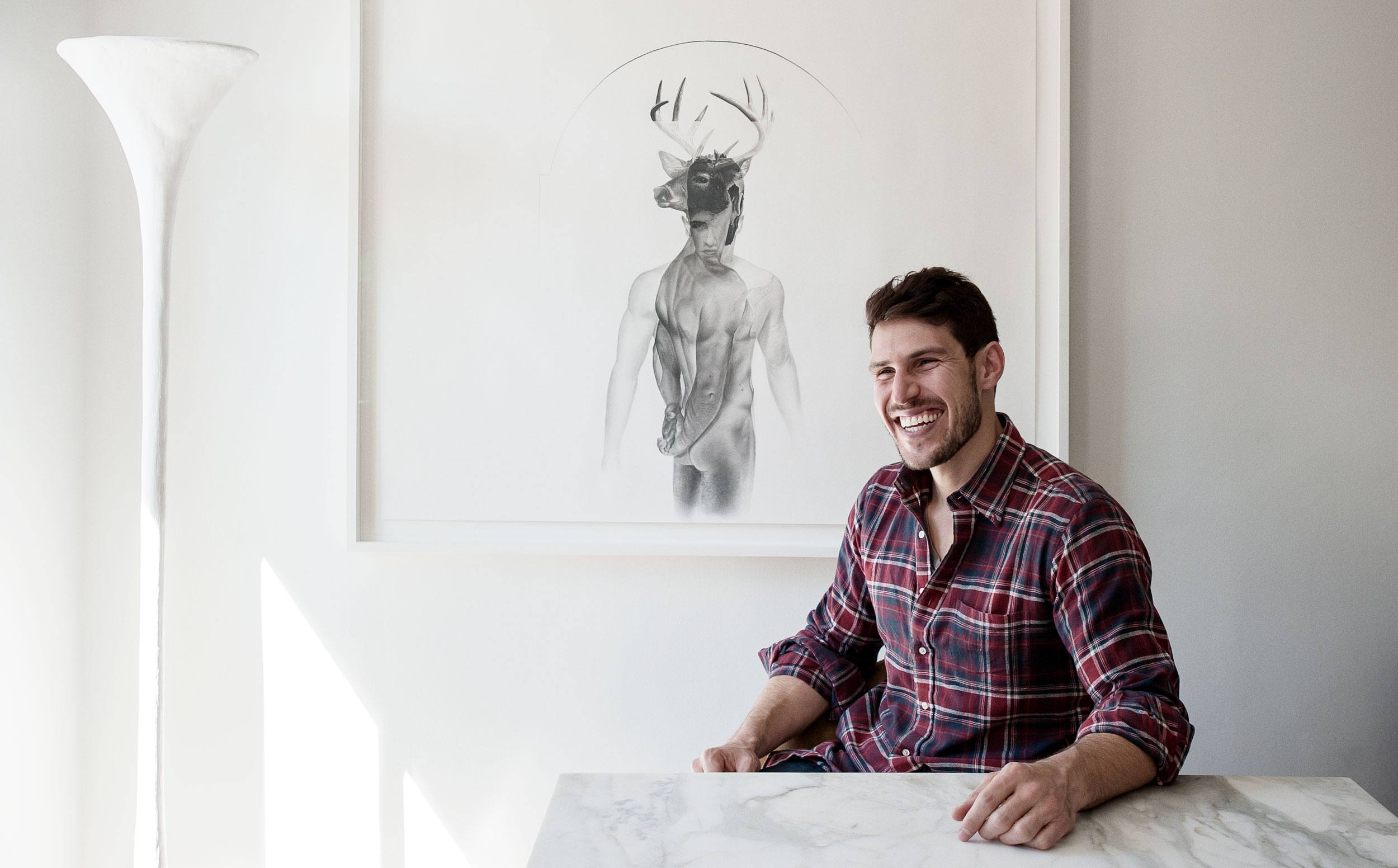 Man in a plaid shirt smiles while sitting at a marble table beside artwork of a human figure with a deer head.