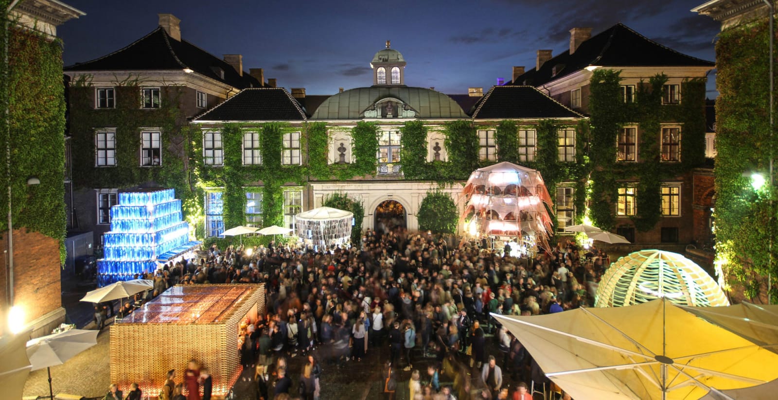 Nighttime festival at an ivy-covered historic building with crowds, colorful lights, and artistic installations.