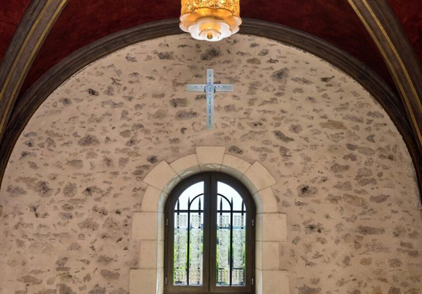 Stone wall with arched window and cross above, under a red vaulted ceiling with a decorative hanging light fixture.