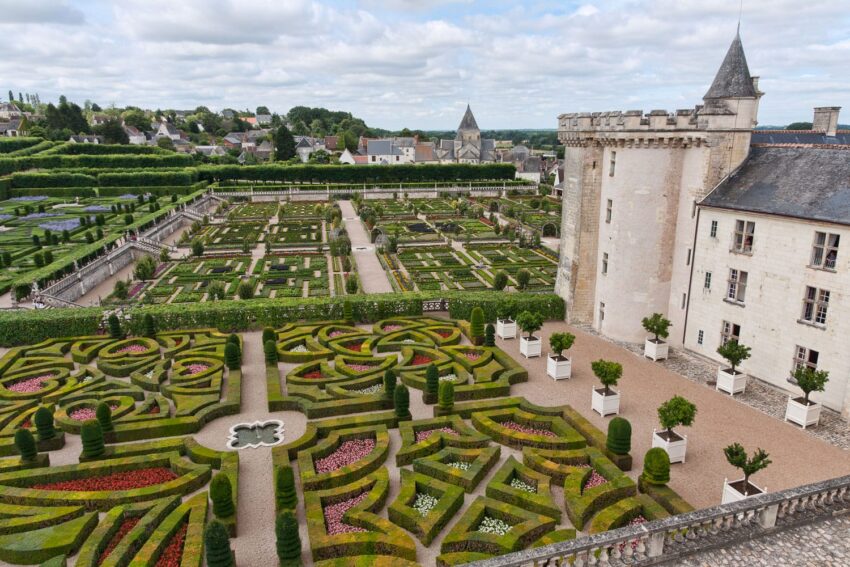 Elaborate formal gardens with geometric patterns beside a historic stone castle and tower under a partly cloudy sky.