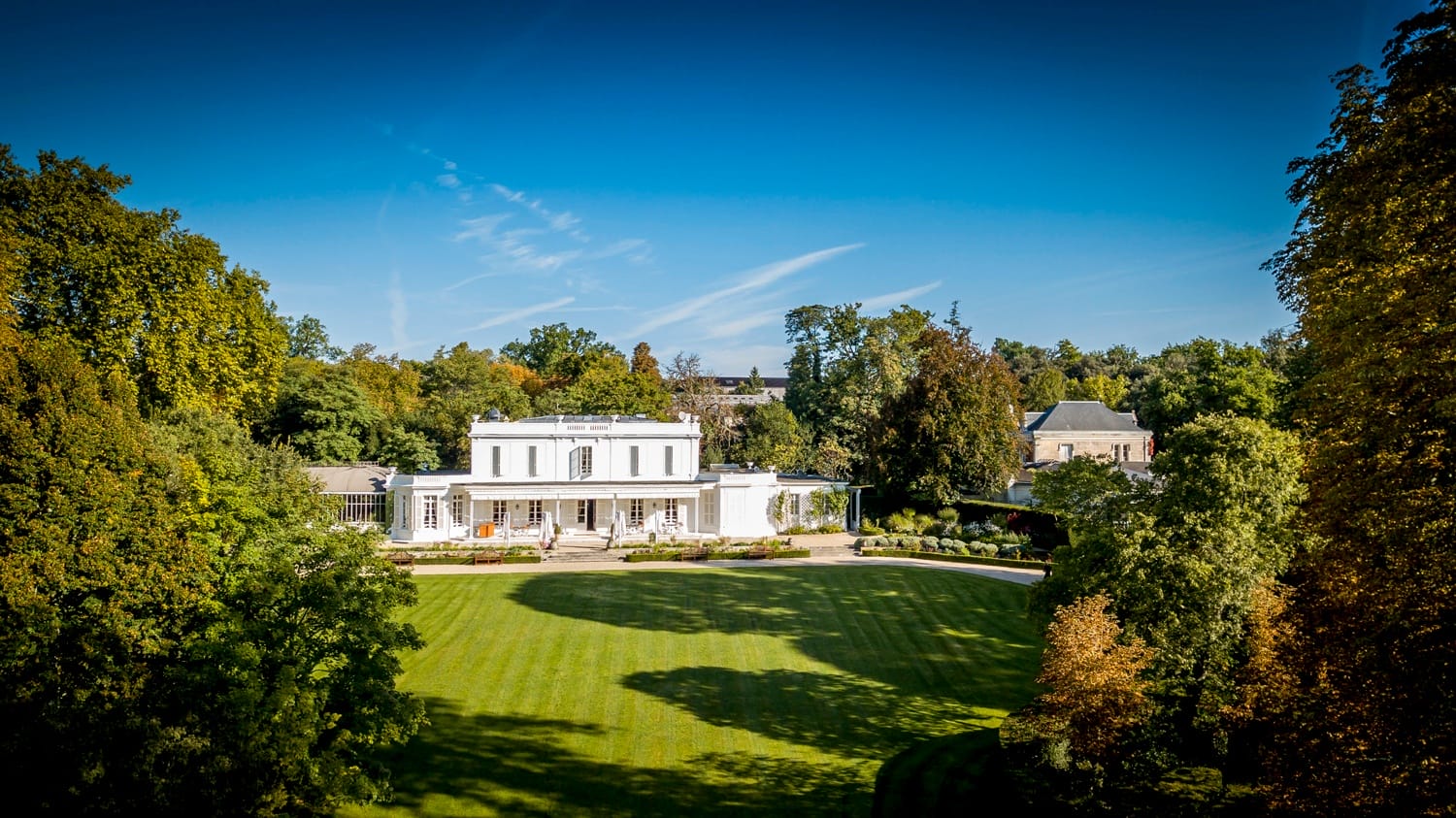 Aerial view of a large white mansion surrounded by trees with a manicured lawn in the foreground under a clear blue sky.