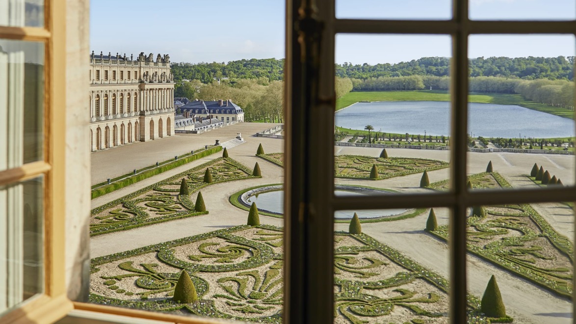 View of ornate gardens and a pond from a window at a historic European estate on a sunny day.