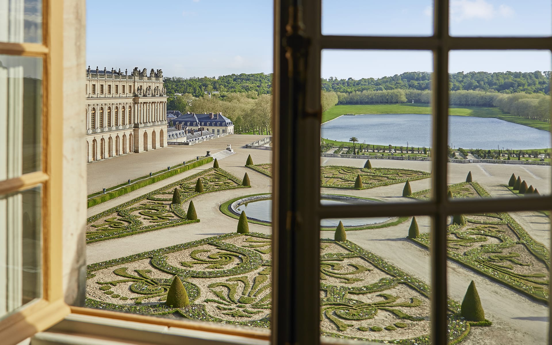 View of ornate gardens and a pond from a window at a historic European estate on a sunny day.