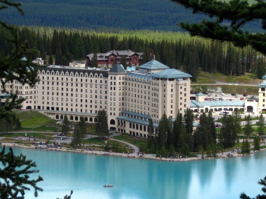 Large lakeside hotel surrounded by trees, with a small boat on the turquoise water in the foreground.