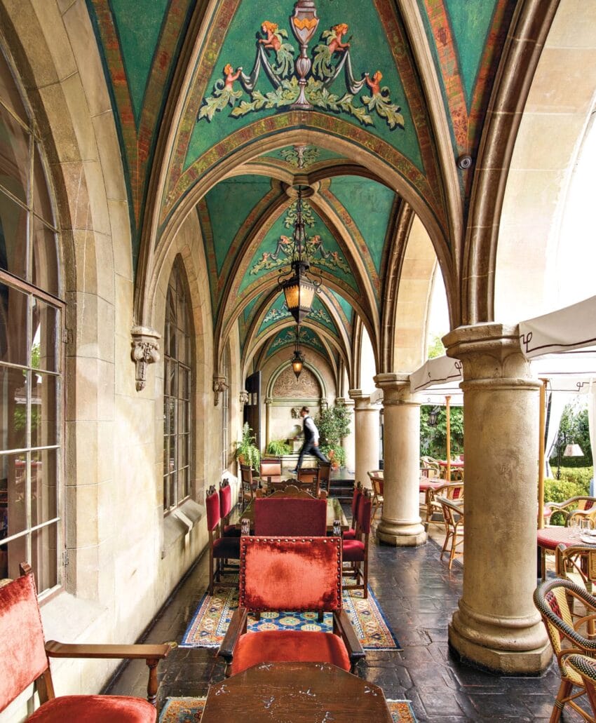 Ornate arched walkway with colorful ceiling design, columns, and red upholstered chairs in a garden cafe setting.