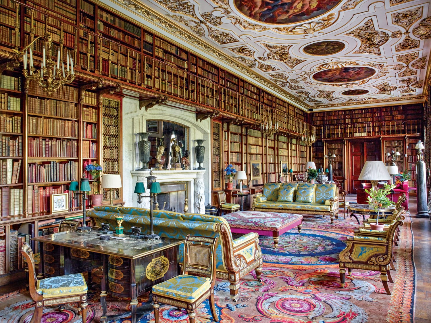 Ornate historic library with a decorative ceiling, plush antique furniture, colorful rugs, and floor-to-ceiling bookshelves.
