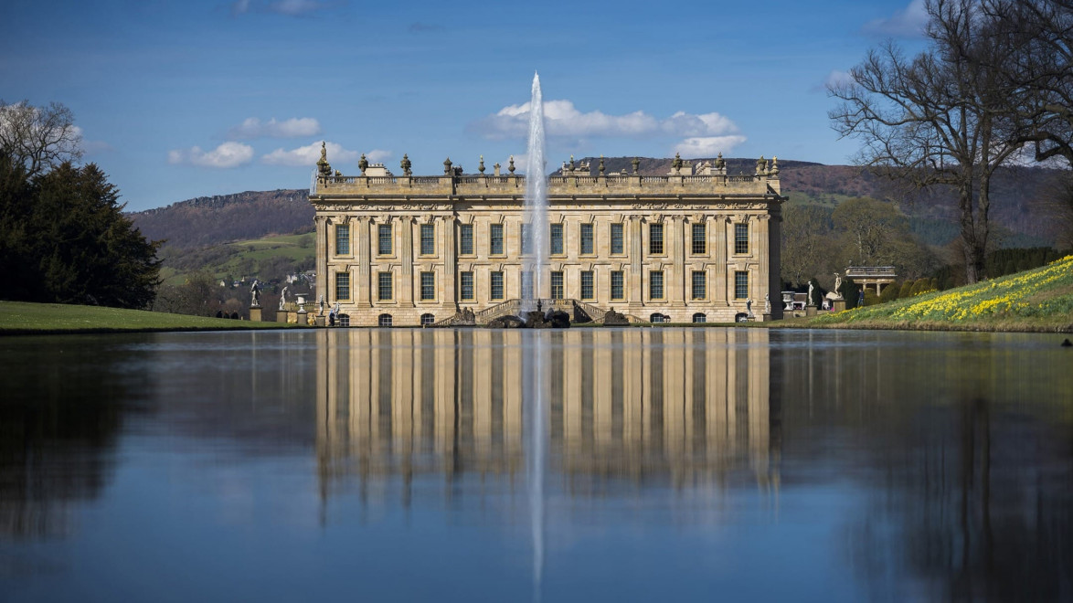 Historic mansion with symmetrical reflection in a calm lake, surrounded by lush lawns and distant hills under a blue sky.