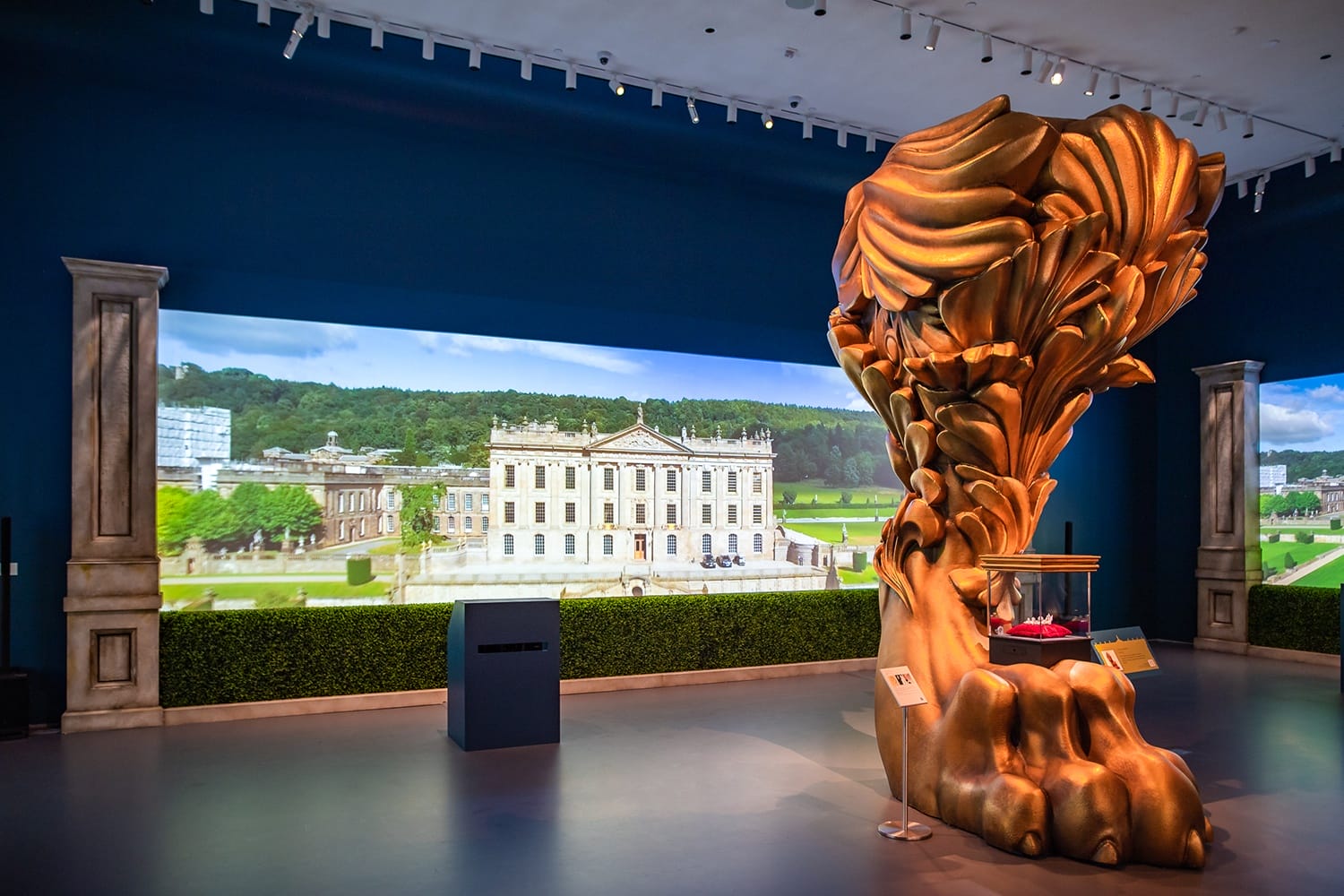 Large golden sculpture inside a modern gallery with a backdrop of a historic estate and lush greenery.