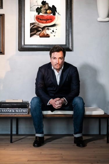 Man seated on a bench in a room with framed artwork on the wall behind him.