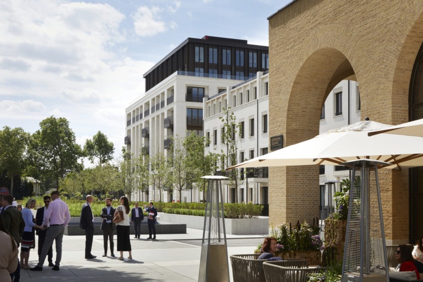 People socializing in an outdoor plaza with modern buildings, archway, and parasol-covered tables under a partly cloudy sky.