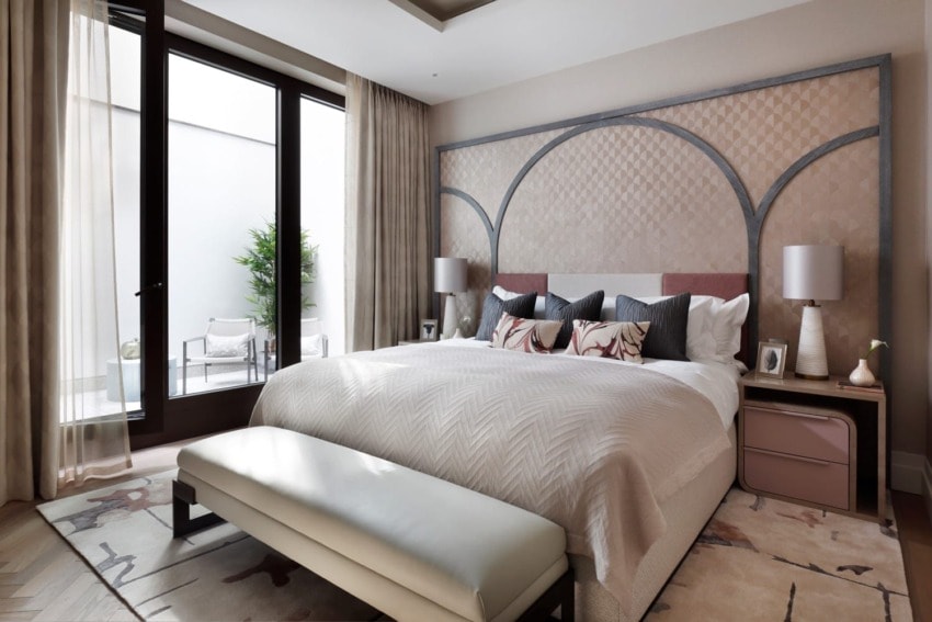 Modern bedroom with large bed, decorative pillows, bench at the foot, and floor-to-ceiling windows leading to a patio.