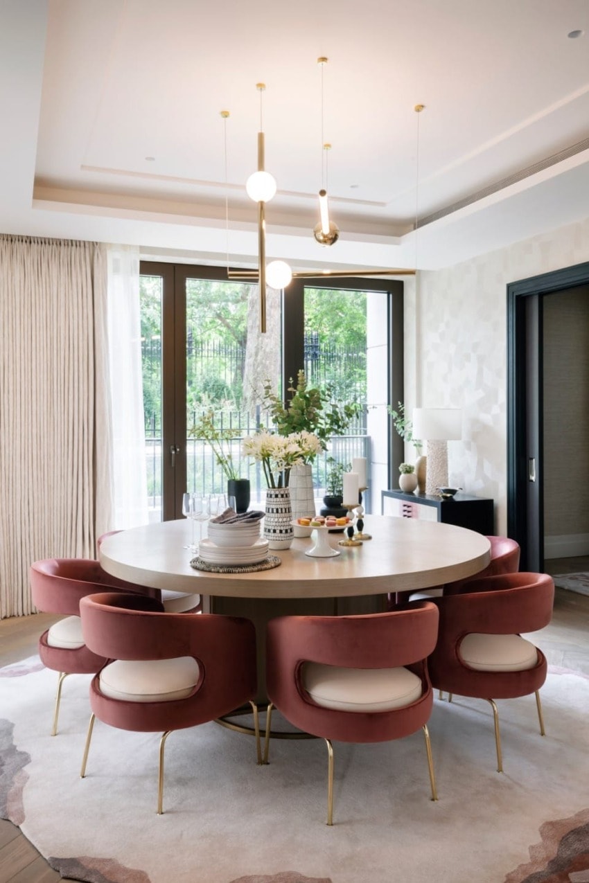 Elegant dining room with round table, pink chairs, modern lighting, large windows, and decorative items on table.