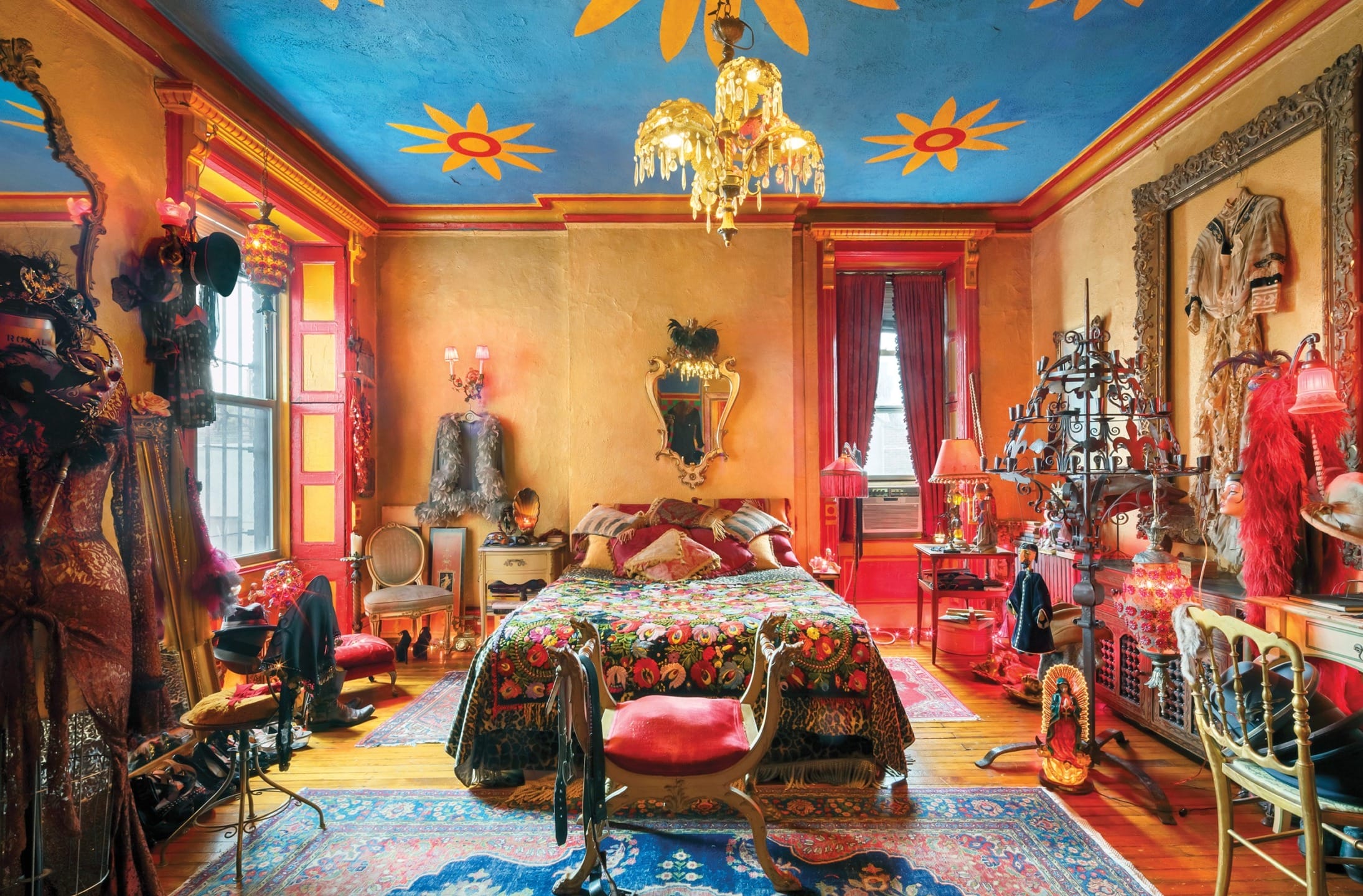Colorful eclectic bedroom with ornate decor, patterned bedding, vibrant ceiling art, chandelier, and vintage furniture.