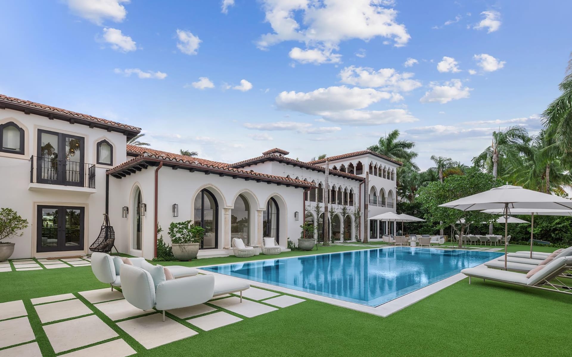 Luxury villa with arches, pool, lounge chairs, and umbrella, surrounded by lush greenery and a clear blue sky.