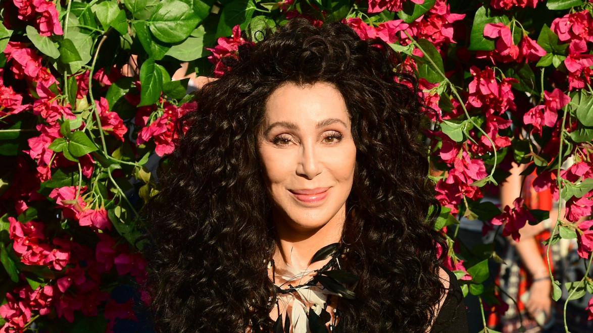 Person with long curly black hair smiling in front of a background full of vibrant pink flowers and green leaves.