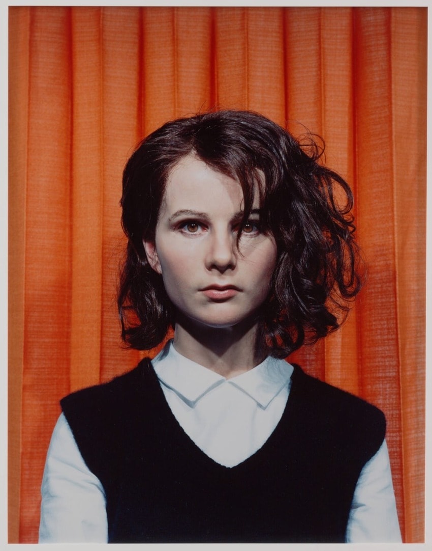 Young woman with shoulder-length hair wearing a black vest and white shirt, standing in front of orange curtains.