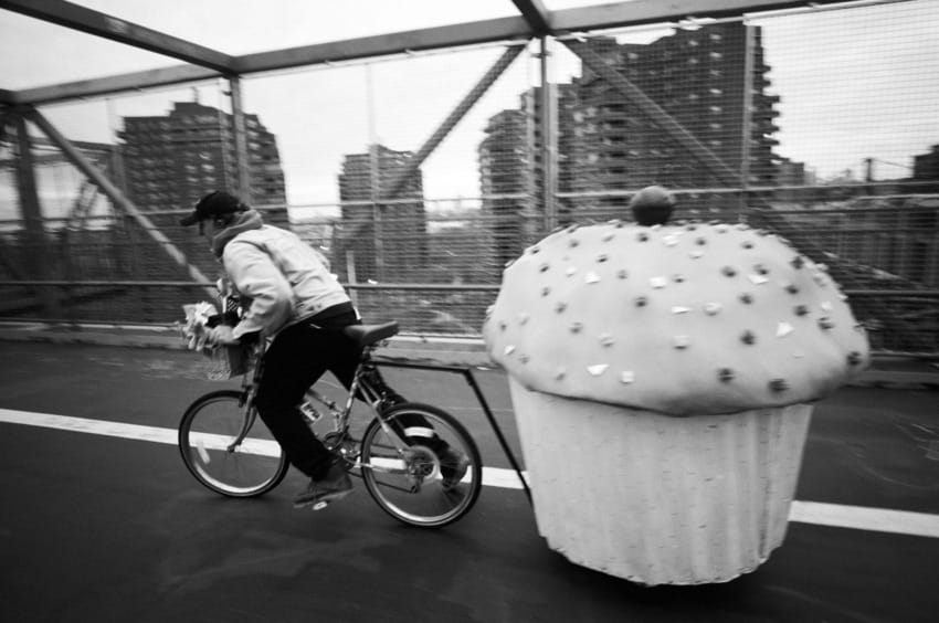 Person biking across a bridge, towing a large cupcake-shaped cart, buildings visible in the background.