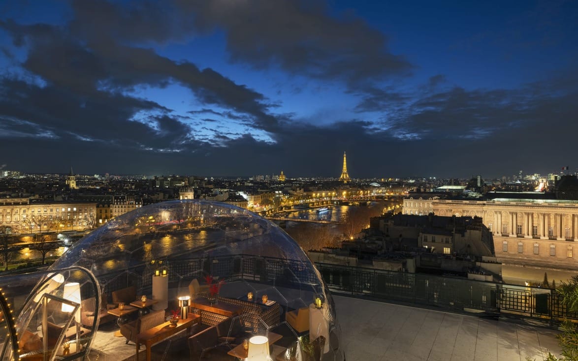 View of Paris skyline at night with Eiffel Tower in the distance and a rooftop dining area under a transparent dome in the foreground.