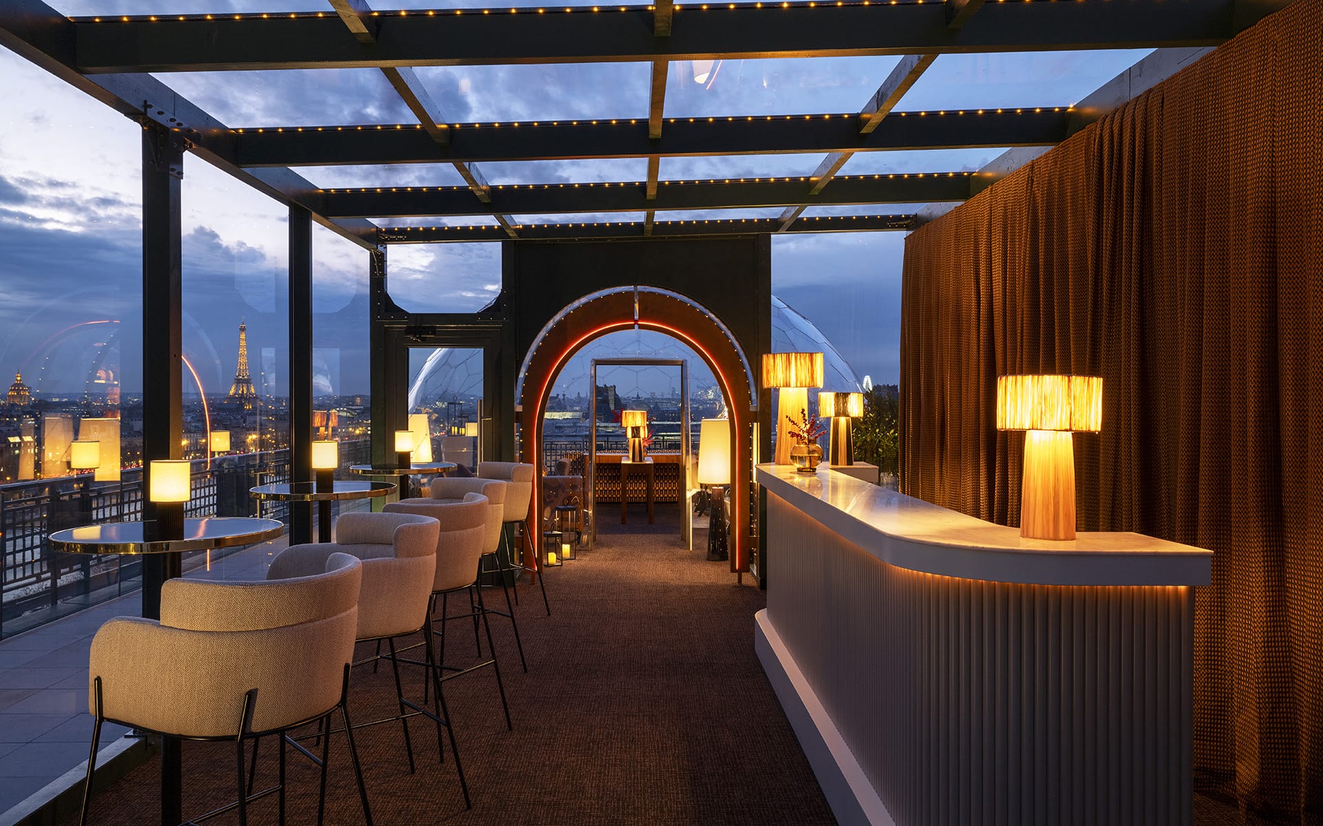 Rooftop bar at dusk with modern seating, illuminated lamps, and city skyline in the background.