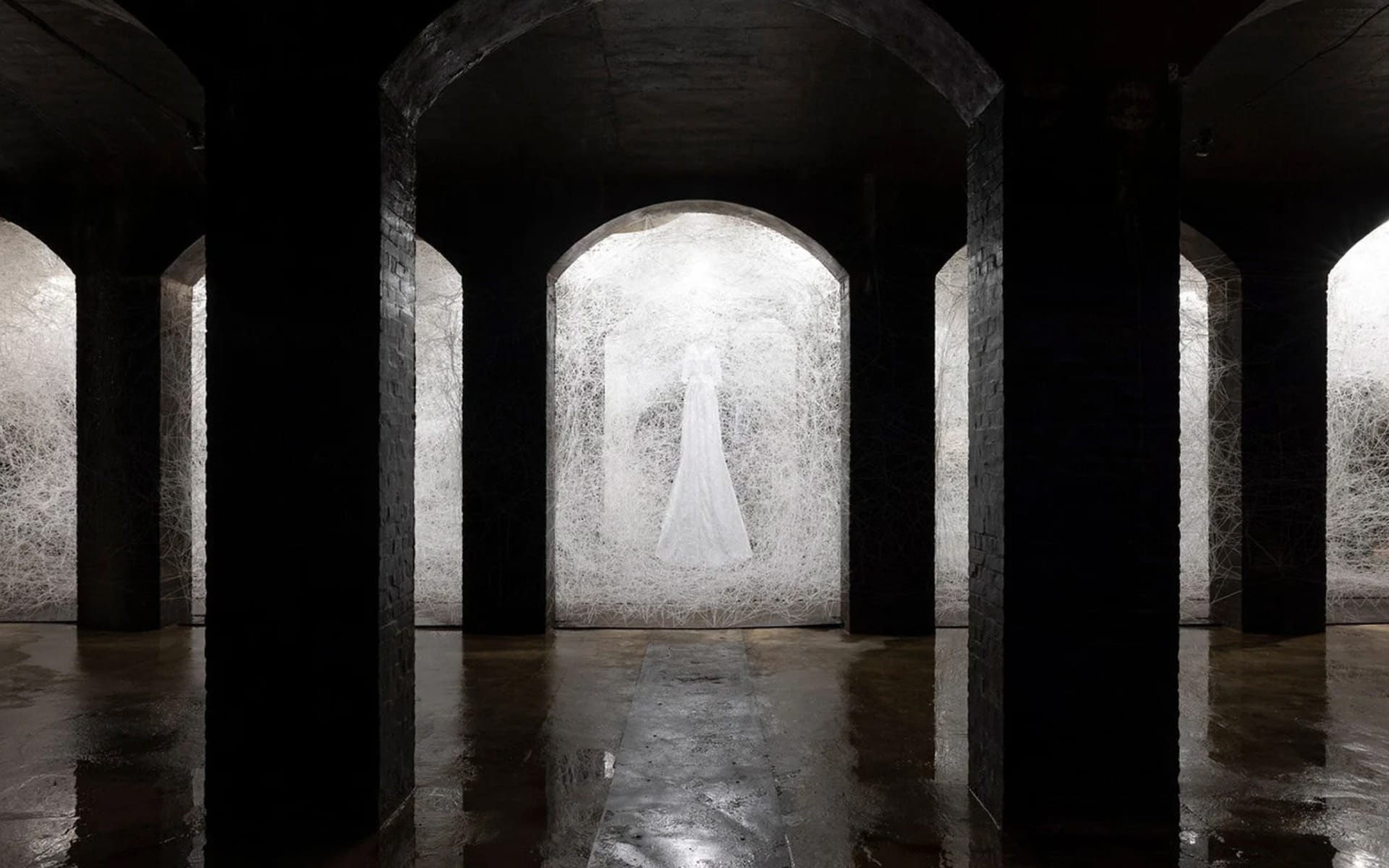 Dark arched hallway with a glowing white dress suspended in spiderweb-like threads creating an ethereal atmosphere.