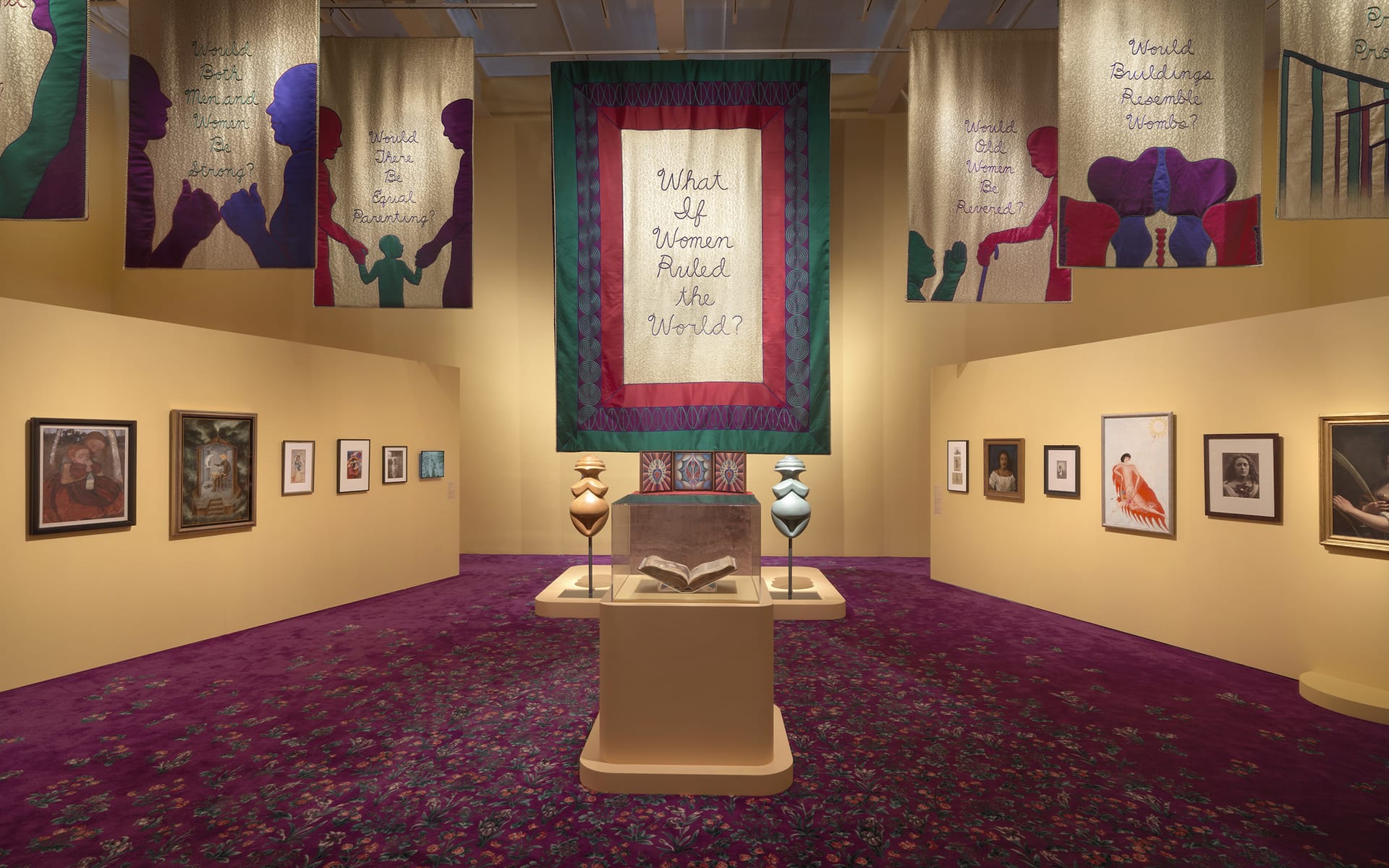 Art exhibition with portraits on walls and a central display with a book, surrounded by tapestries reading "What If Women Ruled the World?"