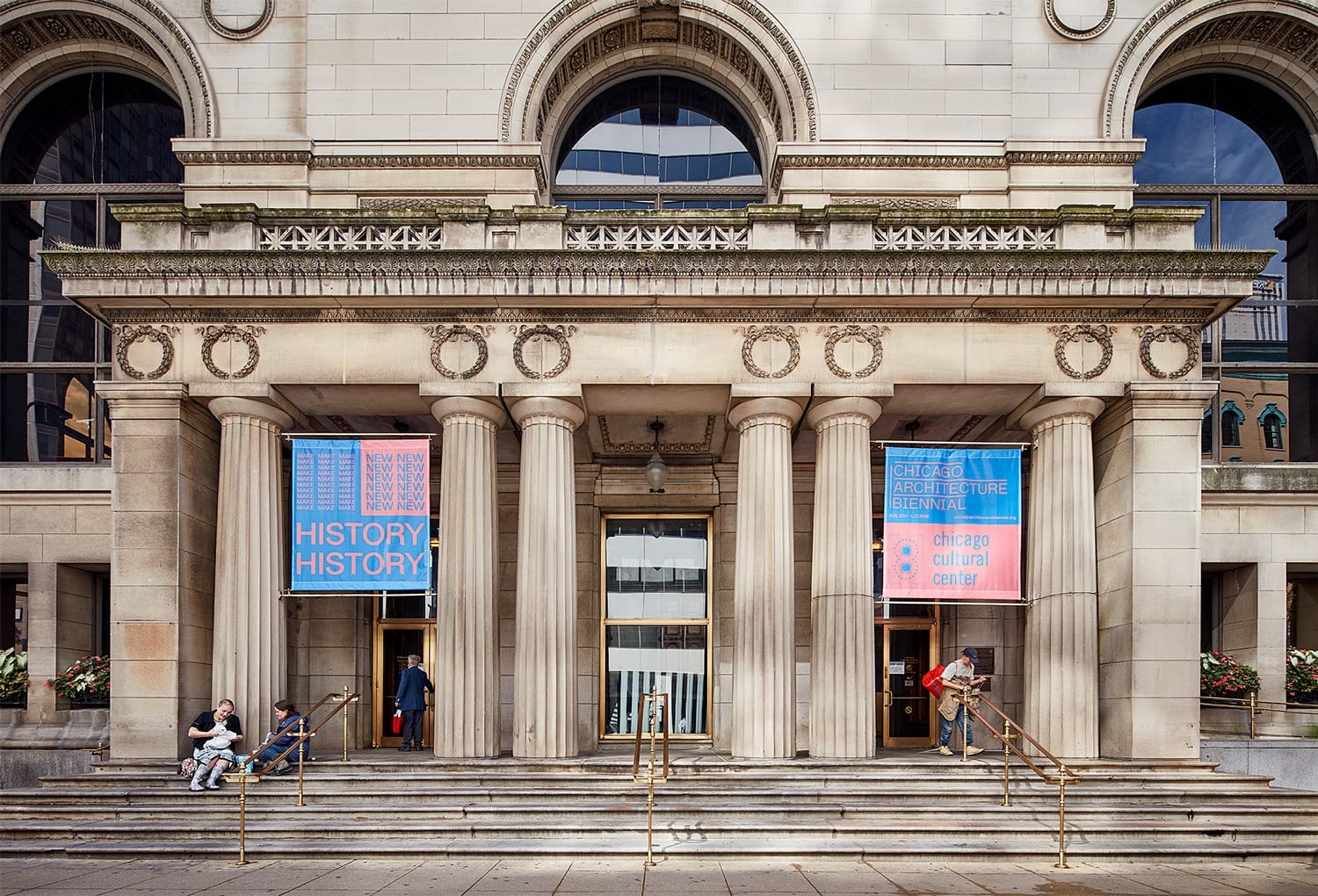 The entrance of a historic building with large columns, colorful banners, and people sitting on the steps.