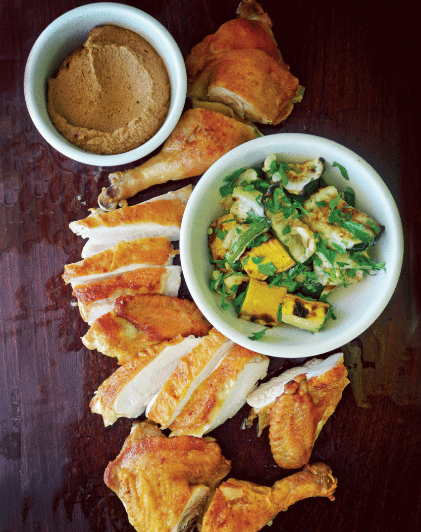 Roasted chicken slices with a bowl of creamy sauce and a dish of zucchini and squash on a dark wooden table.