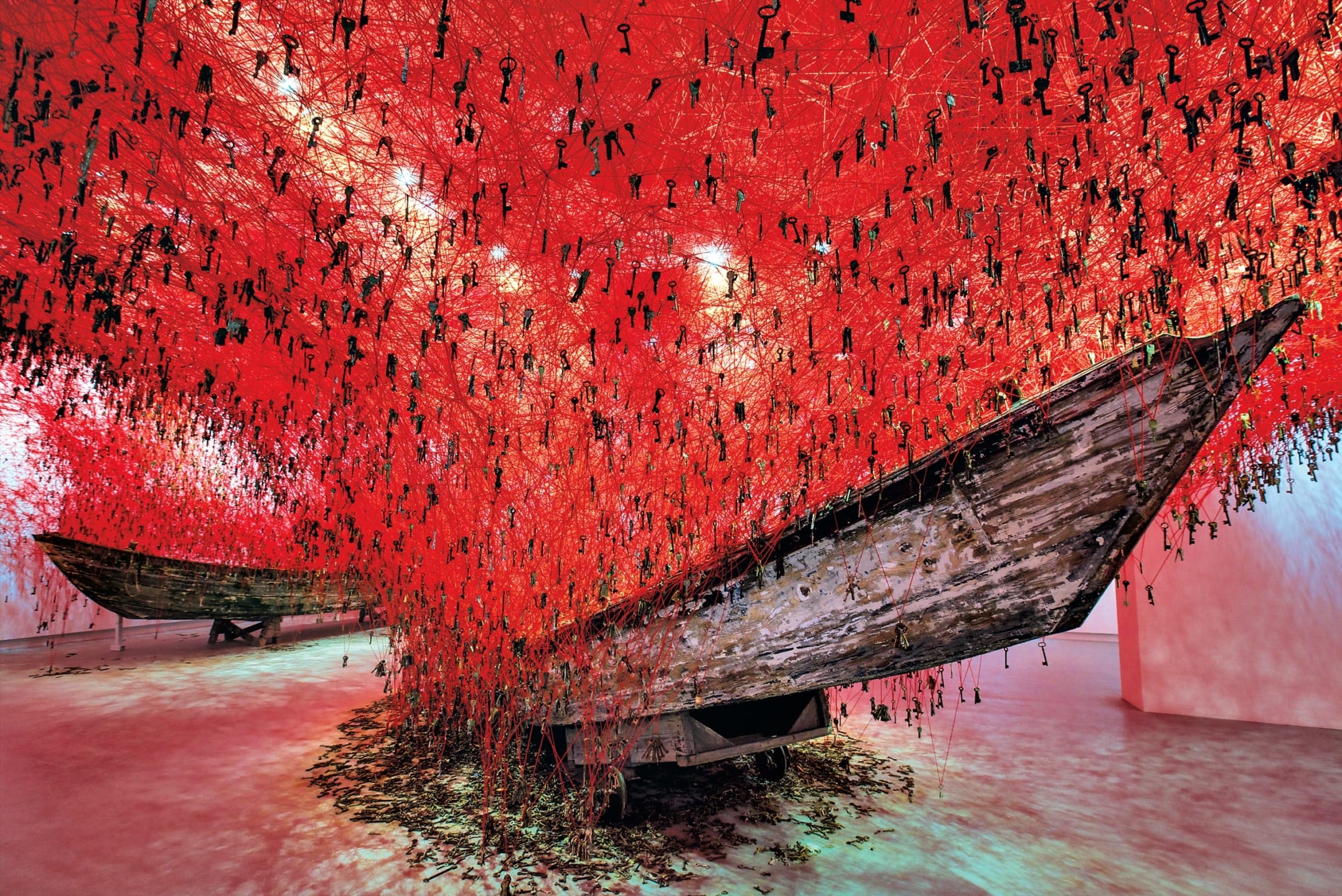 Bright red art installation with suspended keys and wooden boats in a large exhibition space.