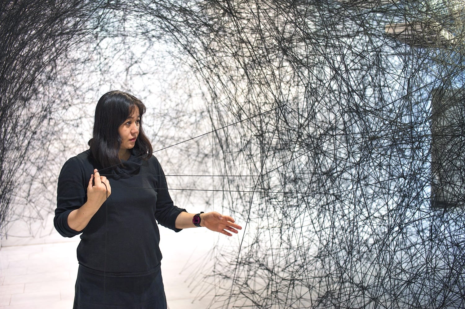 Woman interacting with intricate black thread art installation in a gallery, gesturing and observing the artwork closely.