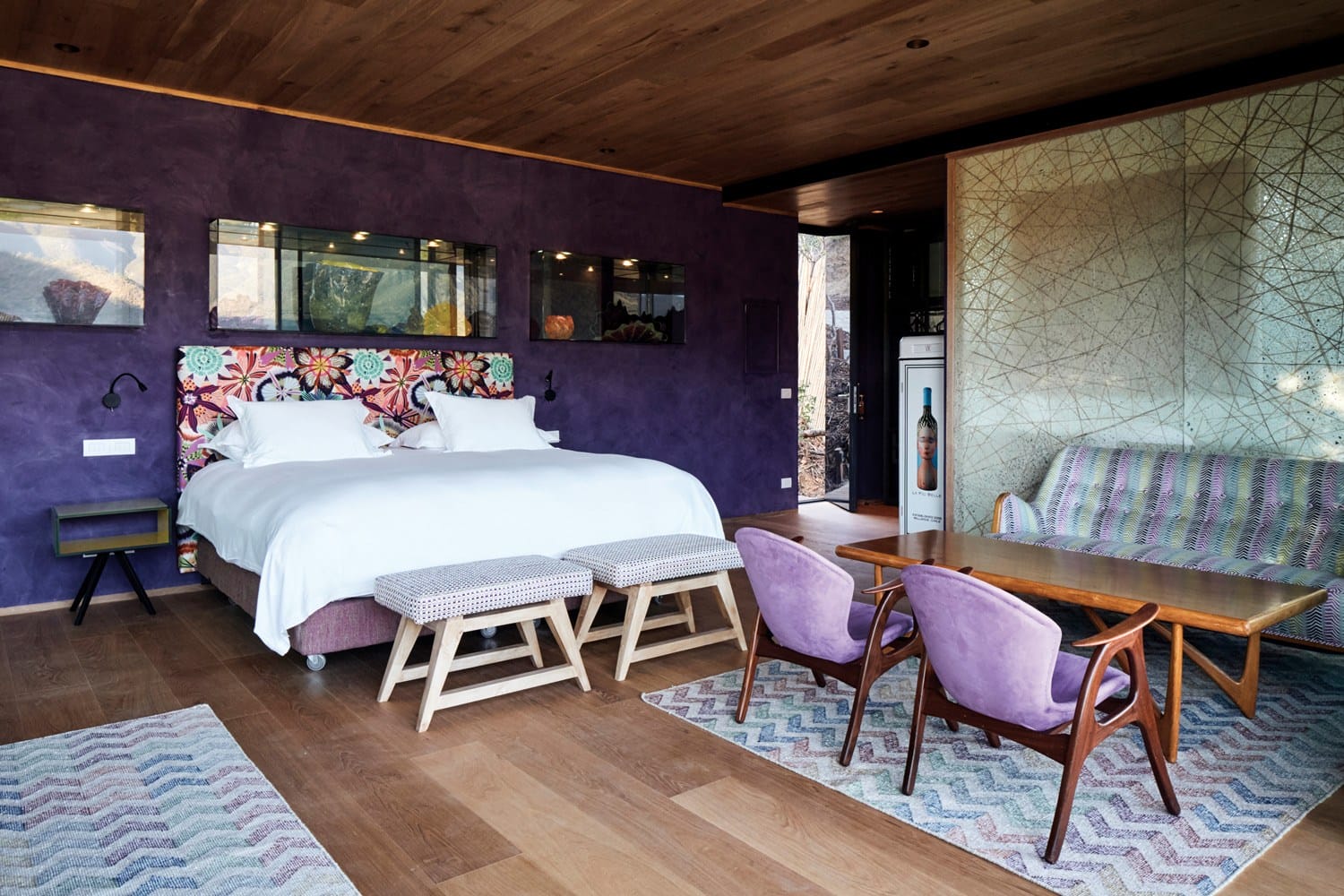 Stylish hotel room with purple walls, patterned bedhead, wooden floor, and modern furniture including a table and chairs.