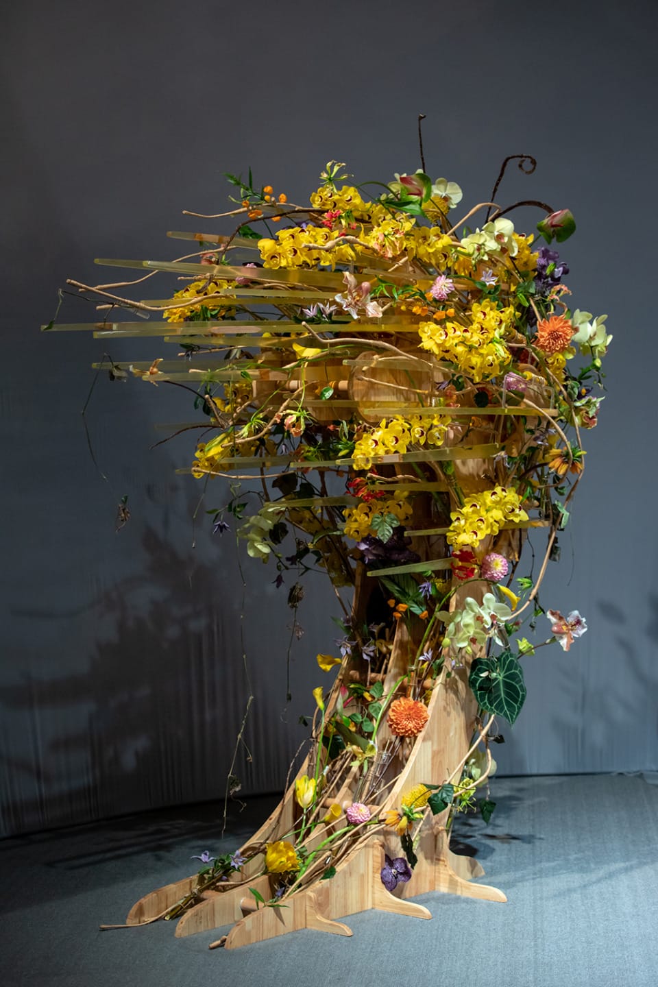 Floral sculpture with vibrant yellow and orange flowers, intertwined branches, and leaves on a wooden stand.